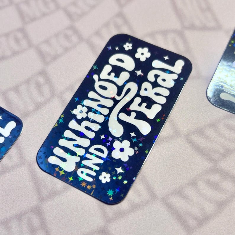 May include: A rectangular sticker with a dark blue background and holographic accents. The sticker features the words "UNHINGED AND FERAL" in white, stylized lettering, along with small white flowers and stars.