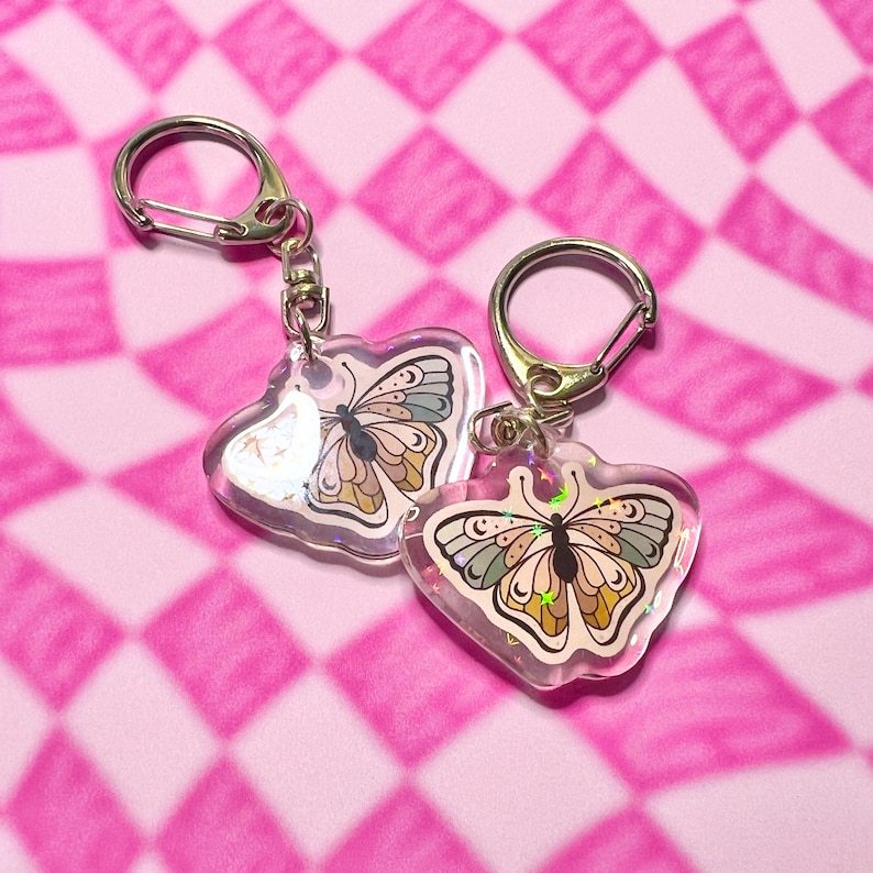 May include: Two clear heart-shaped keychains with silver key rings. Each keychain features a colorful butterfly with iridescent glitter accents.