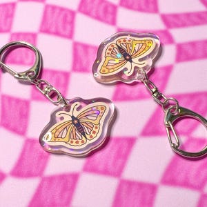 May include: Two iridescent butterfly keychains with silver key rings. The butterflies are yellow, orange, and pink with a black outline and small white flowers.