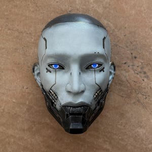 May include: A futuristic, gray and black robotic face with glowing blue eyes. The face has detailed mechanical features, including a segmented lower jaw and lines across the forehead. The head is set against a brown background.