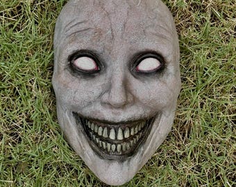 May include: A full-face mask featuring a pale, textured, human-like visage with deep-set, hollow eye sockets containing solid white eyes, and a wide, exaggerated grin revealing a detailed set of stained, realistic teeth and dark, shadowed gums.