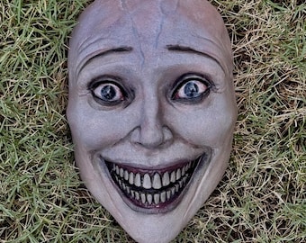 May include: A pale, humanoid mask with a smooth, rounded forehead, prominent brow ridges, and deep-set blue eyes. The face features a wide, exaggerated grin revealing a full set of realistic, detailed teeth and gums, with subtle shading.
