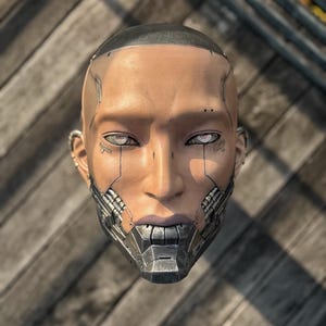 May include: A futuristic, human-like mask with a metallic lower face and detailed circuitry. The mask has a smooth, tan skin tone, with glowing white eyes and a dark, shaved head. The design suggests a cyberpunk or sci-fi theme.