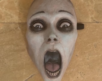 Könnte beinhalten: Eine blasse, menschenähnliche Maske mit weit geöffnetem Mund, dunklen, tief liegenden Augenhöhlen und geschwungenen Augenbrauen. Der Gesichtsausdruck zeigt intensives Entsetzen oder einen Schrei, wobei Zähne und Zunge sichtbar sind.
