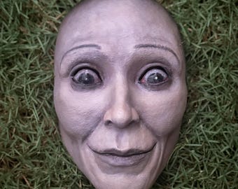 May include: A sculpted mask of a human face with a smooth, pale gray finish. The mask features wide, dark, glossy eyes, arched eyebrows, and a subtle, closed-mouth smile. The facial features are defined with soft shading and realistic contours.