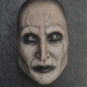 Könnte beinhalten: Eine blasse, kahle Maske mit einem hageren Gesicht, tief liegenden Augen und dunklen Lippen. Die Maske hat sichtbare Adern und Risse mit einem finsteren Ausdruck. Die Augen sind von blassgelb-grüner Farbe, was das unheimliche Aussehen verstärkt. Ein Halloween- oder Horror-Artikel.