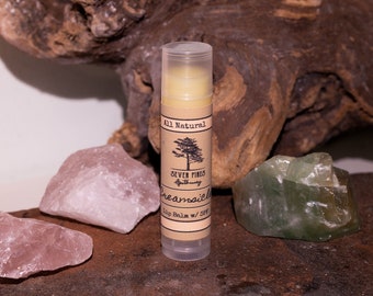 May include: A brown and white tube of lip balm with the text "Seven Pines Apothecary" and "Dreamsicle Lip Balm w/ SPF" on the label. The tube is sitting on a wooden surface with pink and green crystals.