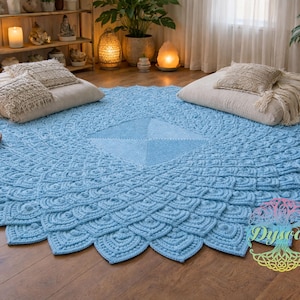 May include: A large, round, light blue crochet rug with a detailed, textured pattern. The rug is placed on a wooden floor, with pillows and candles in the background. The rug's design features a central square and a radiating, fan-like pattern.