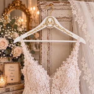 May include: A white wooden clothes hanger featuring a gold-toned metal hook and a central oval medallion with a gold letter M surrounded by small crystals. The hanger is adorned with gold filigree accents on both sides of the central emblem.