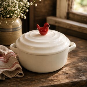 May include: White ceramic casserole dish with a red bird-shaped handle on the lid. The dish has two side handles and is sitting on a wooden surface. A vase of white flowers is in the background.