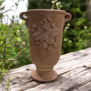 May include: A terracotta-colored vase with a floral relief design. The vase has two small handles and a pedestal base. The floral design features leaves and berries. The vase is suitable for indoor or outdoor use.