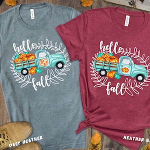 Könnte beinhalten: Zwei T-Shirts mit einem Design eines blauen Pick-up-Trucks, der Kürbisse transportiert, und dem Text "hello fall". Das T-Shirt links ist in der Farbe Deep Heather und das T-Shirt rechts in der Farbe Heather Berry.