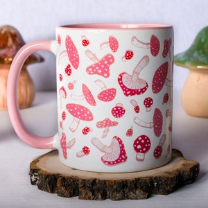 May include: A white ceramic mug with a pink rim and handle. The mug features a repeating pattern of pink and red mushrooms.