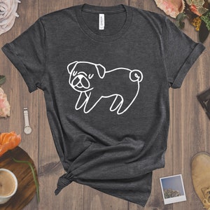 May include: A dark gray t-shirt with a white outline of a pug dog. The pug is facing the left side of the image and has its tongue sticking out.
