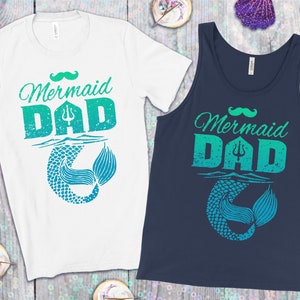 Puede incluir: Dos camisetas con un gráfico desgastado en verde azulado y verde de una cola de sirena y el texto "Mermaid Dad" con un bigote sobre el texto. Una camiseta es una camiseta blanca y la otra es un tanque azul marino.
