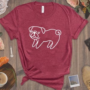May include: A maroon t-shirt with a white outline of a pug dog. The shirt is made of a soft, comfortable material.