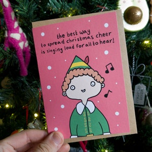 May include: A pink Christmas card featuring a cartoon elf wearing a green and yellow hat. The card reads "the best way to spread christmas cheer is singing loud for all to hear!"