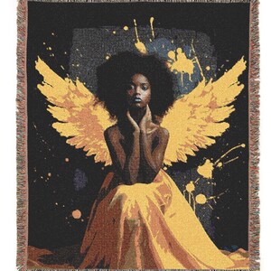 May include: A woven throw blanket featuring an artistic depiction of a person with large yellow wings, seated in a flowing yellow gown against a black background with yellow paint splatters. The blanket has a fringed edge.