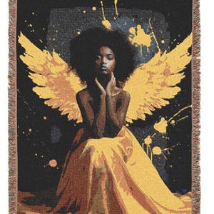May include: Woven throw blanket featuring a woman with yellow angel wings and a yellow dress. The artwork has a black background with yellow paint splatters. The blanket has a fringed edge.