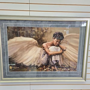 May include: Framed art print featuring a woman with wings sitting with her head down. The woman is wearing a white dress and has dark skin. The background is a soft brown color with a blurred image of a room with framed pictures.