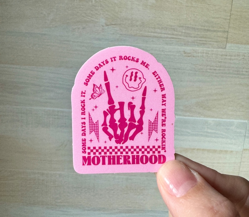 Pu&ograve; includere: Adesivo rosa con la scritta "SOME DAYS I ROCK IT. SOME DAYS IT ROCKS ME. EITHER WAY WE'RE ROCKIN' MOTHERHOOD." Presenta un gesto della mano rock, una farfalla, una faccina sorridente e dei fulmini.