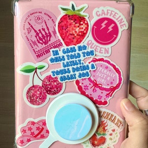 Pu&ograve; includere: Una custodia rosa decorata con vari adesivi. Gli adesivi includono una fragola, ciliegie, un cappello da cowboy e il testo che dice "CAFFEINE QUEEN", "IN CASE NO ONE TOLD YOU LATELY, YOU'RE DOING A GREAT JOB" e "LITERALLY JUST A GIRL". &Egrave; attaccato un supporto per telefono azzurro.