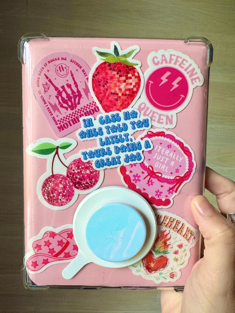 Pu&ograve; includere: Una custodia rosa decorata con vari adesivi. Gli adesivi includono una fragola, ciliegie, un cappello da cowboy e il testo che dice "CAFFEINE QUEEN", "IN CASE NO ONE TOLD YOU LATELY, YOU'RE DOING A GREAT JOB" e "LITERALLY JUST A GIRL". &Egrave; attaccato un supporto per telefono azzurro.