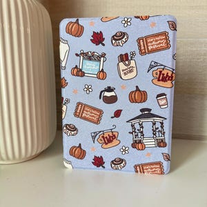 May include: A light blue e-reader cover featuring a pattern of pumpkins, autumn leaves, and the text "Stars Hollow Autumn Festival." Other illustrations include a coffee cup, a gazebo, and the text "Luke's."