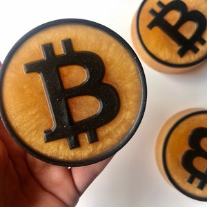 Puede incluir: Tres jabones redondos de color dorado con un símbolo de Bitcoin negro en cada uno. Los jabones están sobre una superficie blanca.