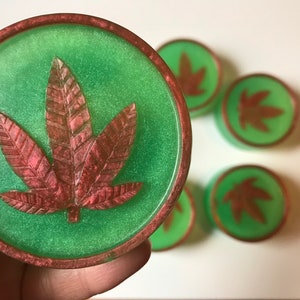 Puede incluir: Un juego de seis posavasos verdes y cobrizos con un diseño de hoja de marihuana.
