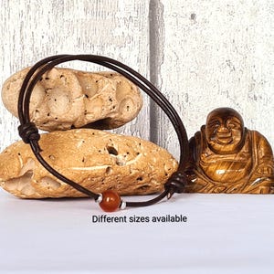 May include: A brown leather bracelet with a red bead and silver details. The bracelet is shown on a white surface with two beige stones and a wooden Buddha statue. The text "Different sizes available" is below the bracelet.