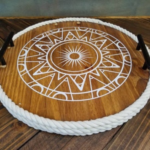 May include: A round wooden tray with a white rope edge and black metal handles. The tray features a white geometric sun design in the centre.