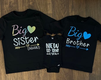 May include: Three black t-shirts with glitter text. The largest shirt says "Big Sister (Again)" with a pink and yellow heart. The medium shirt says "Big Brother" with a blue heart. The smallest shirt says "New to the Crew" with a white heart.