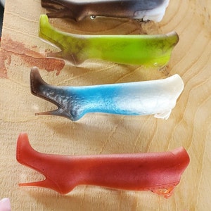 May include: A set of eight different colored resin boot charms. The charms are arranged in a row on a wooden surface. The charms are all in the shape of a boot and are made of a translucent resin. The charms are all different colors, including black, gold, brown, green, blue, red, and brown.