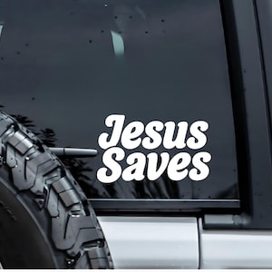 May include: White vinyl decal with the words "Jesus Saves" in a bold, retro font. The decal is affixed to the rear window of a vehicle, with a spare tire visible on the left side. The background is a dark, reflective surface.