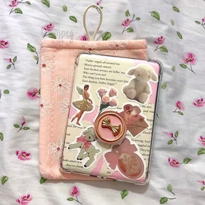 May include: A pink phone case with a clear back, decorated with stickers of a ballerina, a lamb, and roses. A gold bow phone grip is attached. A matching pink pouch with daisy embroidery is next to the phone. The background is a floral pattern.