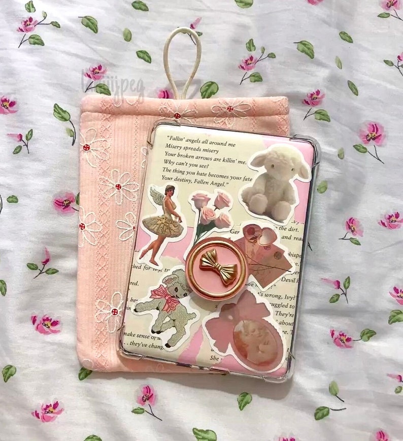 May include: A pink phone case with a clear back, decorated with stickers of a ballerina, a lamb, and roses. A gold bow phone grip is attached. A matching pink pouch with daisy embroidery is next to the phone. The background is a floral pattern.