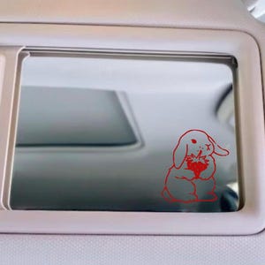 May include: A red line drawing of a rabbit holding a strawberry is on a car's rearview mirror.