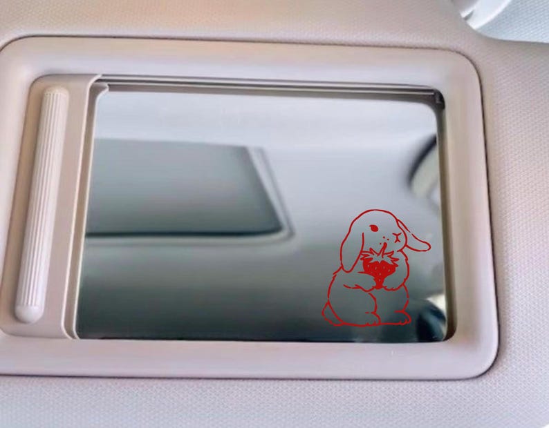 May include: A red line drawing of a rabbit holding a strawberry is on a car's rearview mirror.