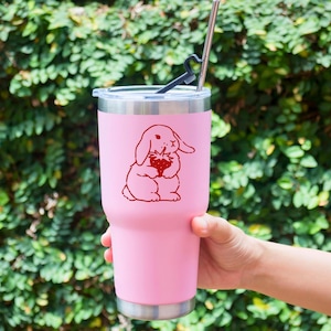 May include: Pink stainless steel tumbler with a red cartoon bunny holding a strawberry. The tumbler has a black lid and a straw.