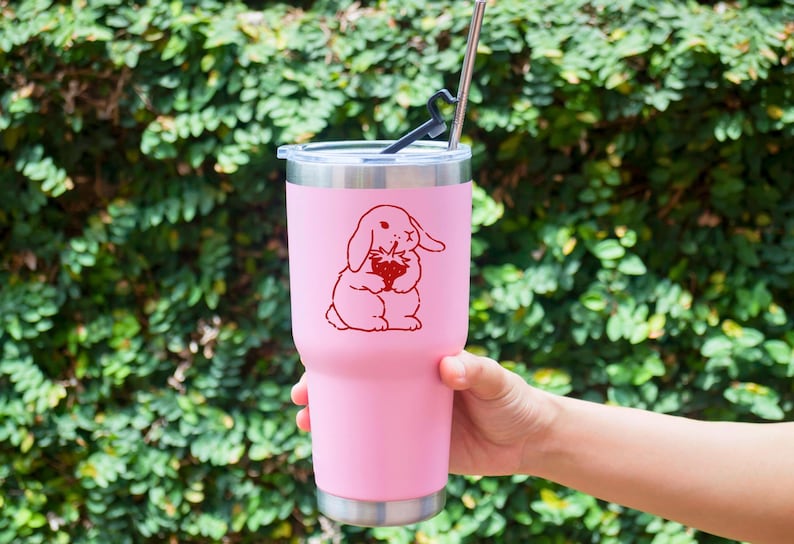 May include: Pink stainless steel tumbler with a red cartoon bunny holding a strawberry. The tumbler has a black lid and a straw.