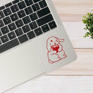 May include: A white laptop computer with a red bunny rabbit sticker on the lid. The bunny is holding a red strawberry. The laptop is on a light wood surface.