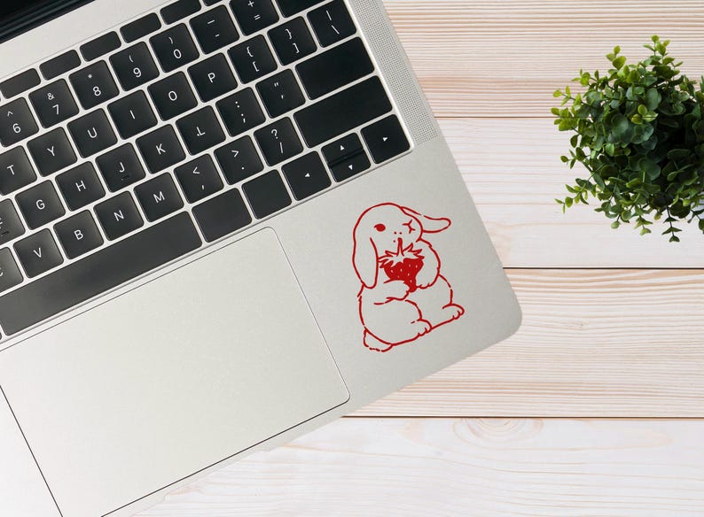May include: A white laptop computer with a red bunny rabbit sticker on the lid. The bunny is holding a red strawberry. The laptop is on a light wood surface.