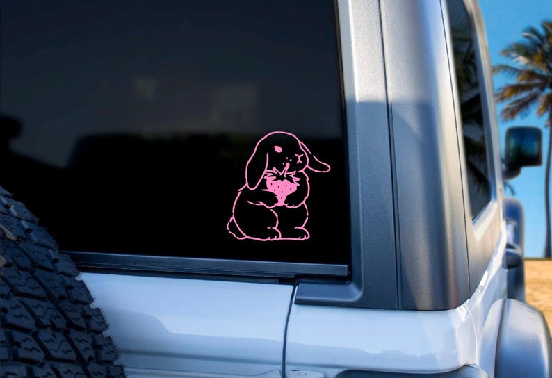May include: Pink vinyl decal of a rabbit holding a strawberry. The rabbit is outlined in black and the strawberry is filled in pink. The decal is on a black car window.