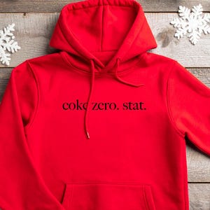 May include: A bright red hoodie with a front pocket and drawstring hood. The text "coke zero, stat." is printed in black across the chest. The hoodie is displayed on a wooden surface with Christmas decorations.