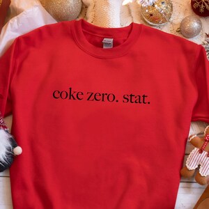 May include: A red sweatshirt with the text "coke zero. stat." in black. The sweatshirt is surrounded by Christmas decorations, including a gift box, ornaments, and a gingerbread man.