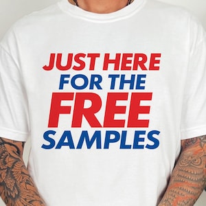 May include: White t-shirt with the text "JUST HERE FOR THE FREE SAMPLES" in red and blue. The shirt is a casual style, suitable for everyday wear. The image shows the shirt on a person.