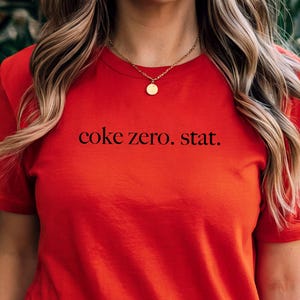 May include: A red t-shirt featuring the black text "coke zero. stat.". The short-sleeved top has a round neck. The image focuses on the upper body of a person wearing the shirt.