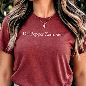 May include: A maroon t-shirt with the text "Dr. Pepper Zero. stat." in white. The shirt is paired with blue jeans. The word "CARDINAL" is in a maroon circle at the bottom right.