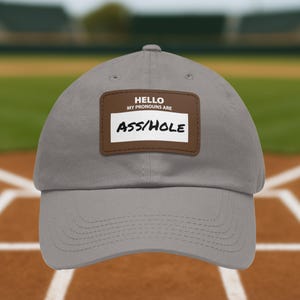 Könnte beinhalten: Graue Baseballkappe mit gebogenem Schirm. Die Kappe hat einen braunen Aufnäher mit dem weißen Text "HELLO MY PRONOUNS ARE ASS/HOLE". Die Kappe besteht aus einem weichen Material und hat eine abgerundete Krone.